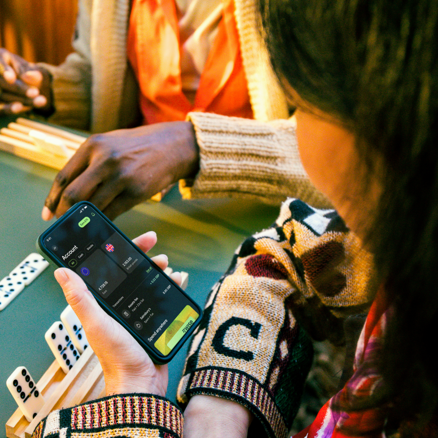 A woman holds a phone while sat at a table. We view the phone from behind her left shoulder, and can see some dominoes on the table beyond.