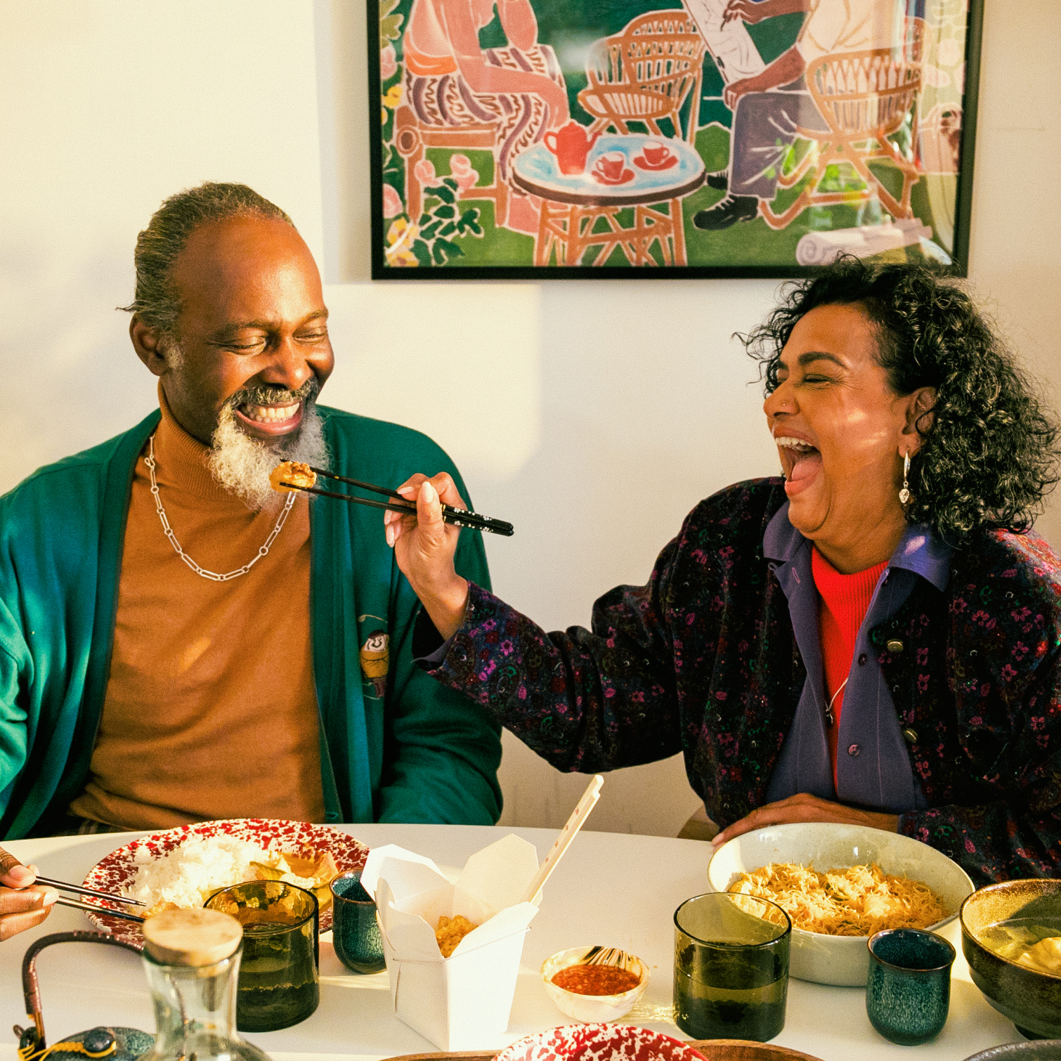 Two people sit at a dining table, with a takeaway meal in front of them. The woman laughs as she holds a bit of food with her chopsticks, and lifts it up towards the man's mouth. 