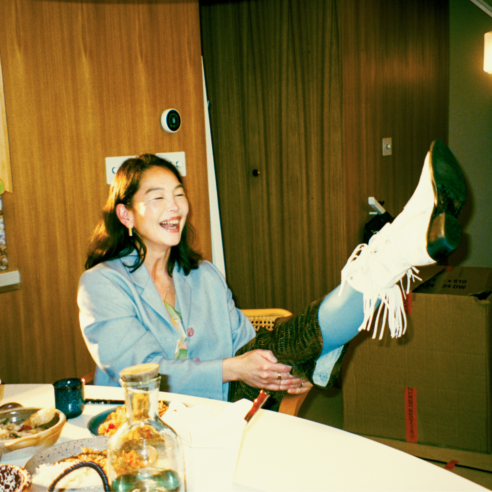 A woman laughing while sat at a dining table, kicking her left leg up to show her fringed cowboy boot.