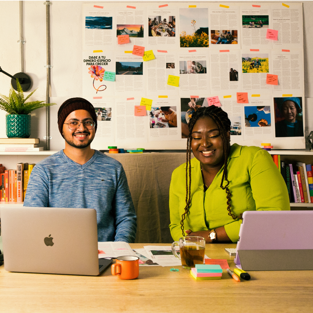 Two people sat at a desk, facing the camera, with their laptop in front of them, and a wall of paper and notes behind them.