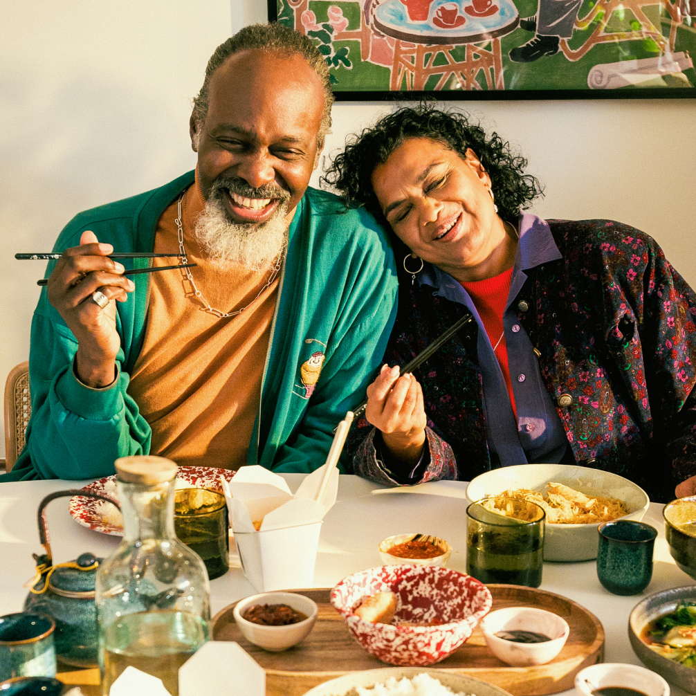 Two people sat smiling at a dining table  that's full of food. The main holds his chopsticks up in front of him, while the woman leans gently on his shoulder.