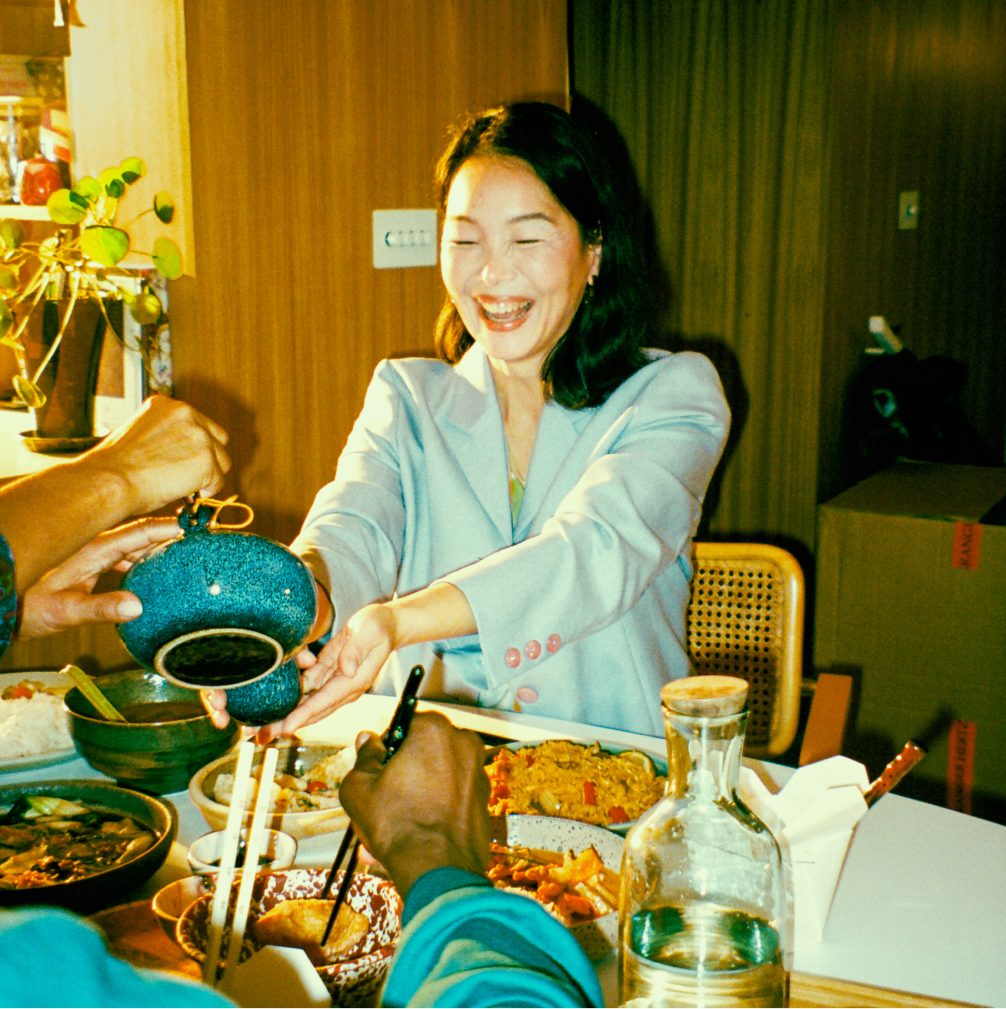 A woman sits smiling at a dinner table, holding out her cup while someone pours her some tea.