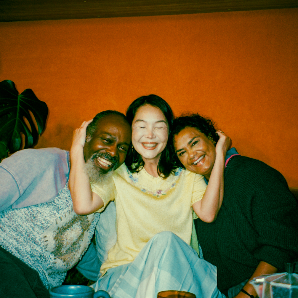 Three people sat together smiling. The woman in the centres gently pulls the two towards her, with her arms cradling each of their heads.