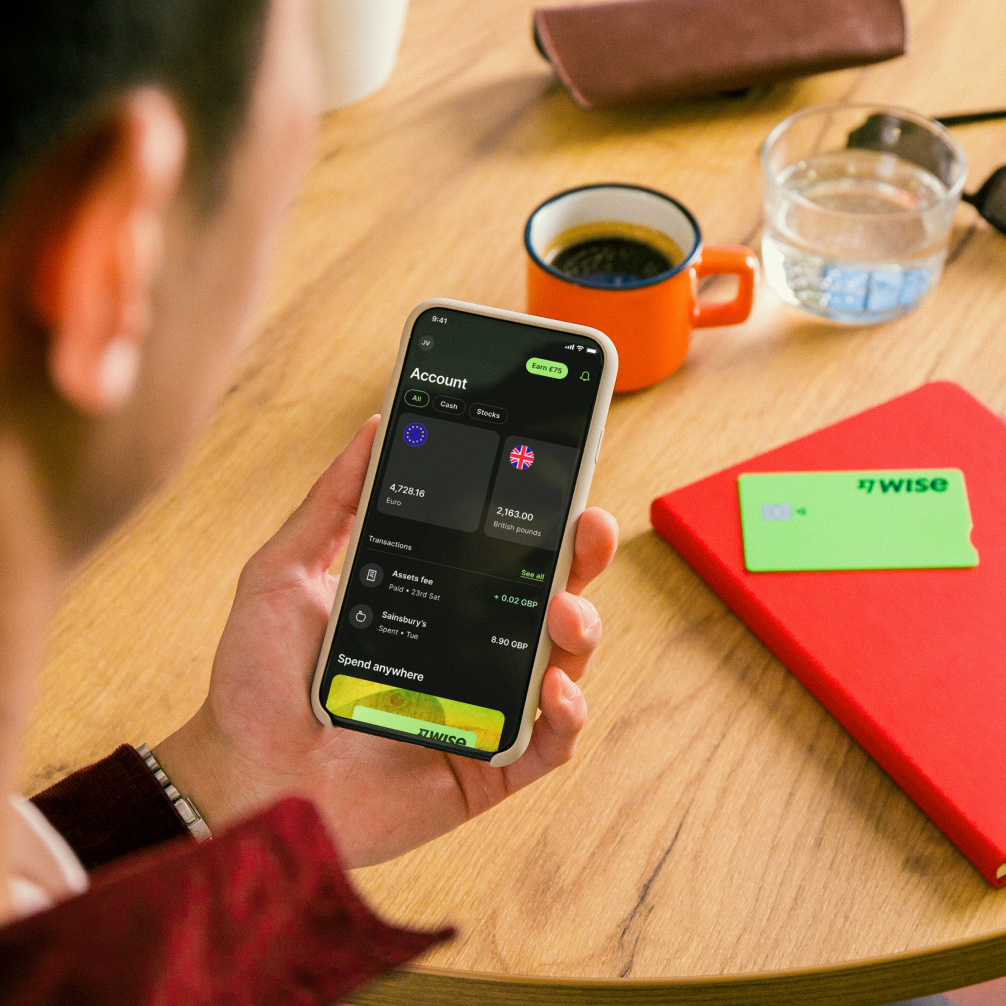 A man holding a phone showing the Wise app, with a Wise debit card pictured on the table in front of him.