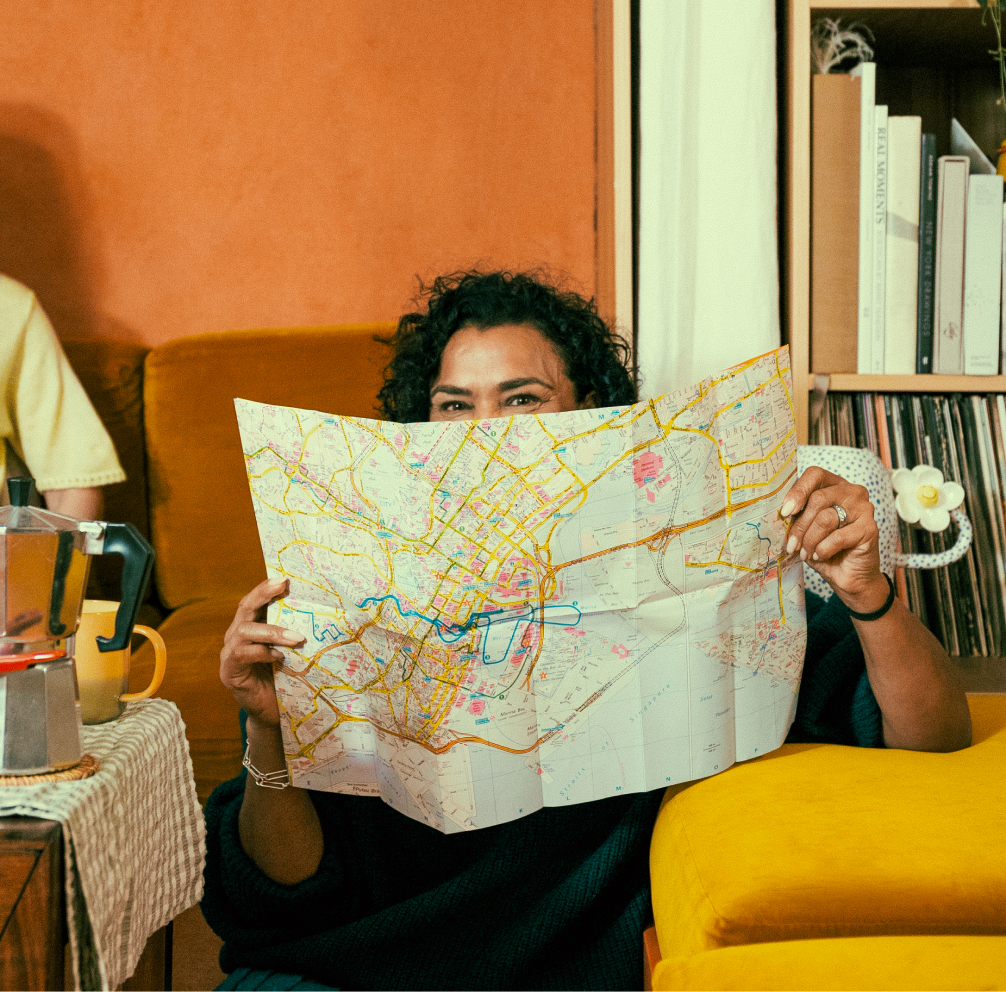 A woman sits smiling on a living room floor, facing the camera, holding up a map to obscure the lower half of her face.