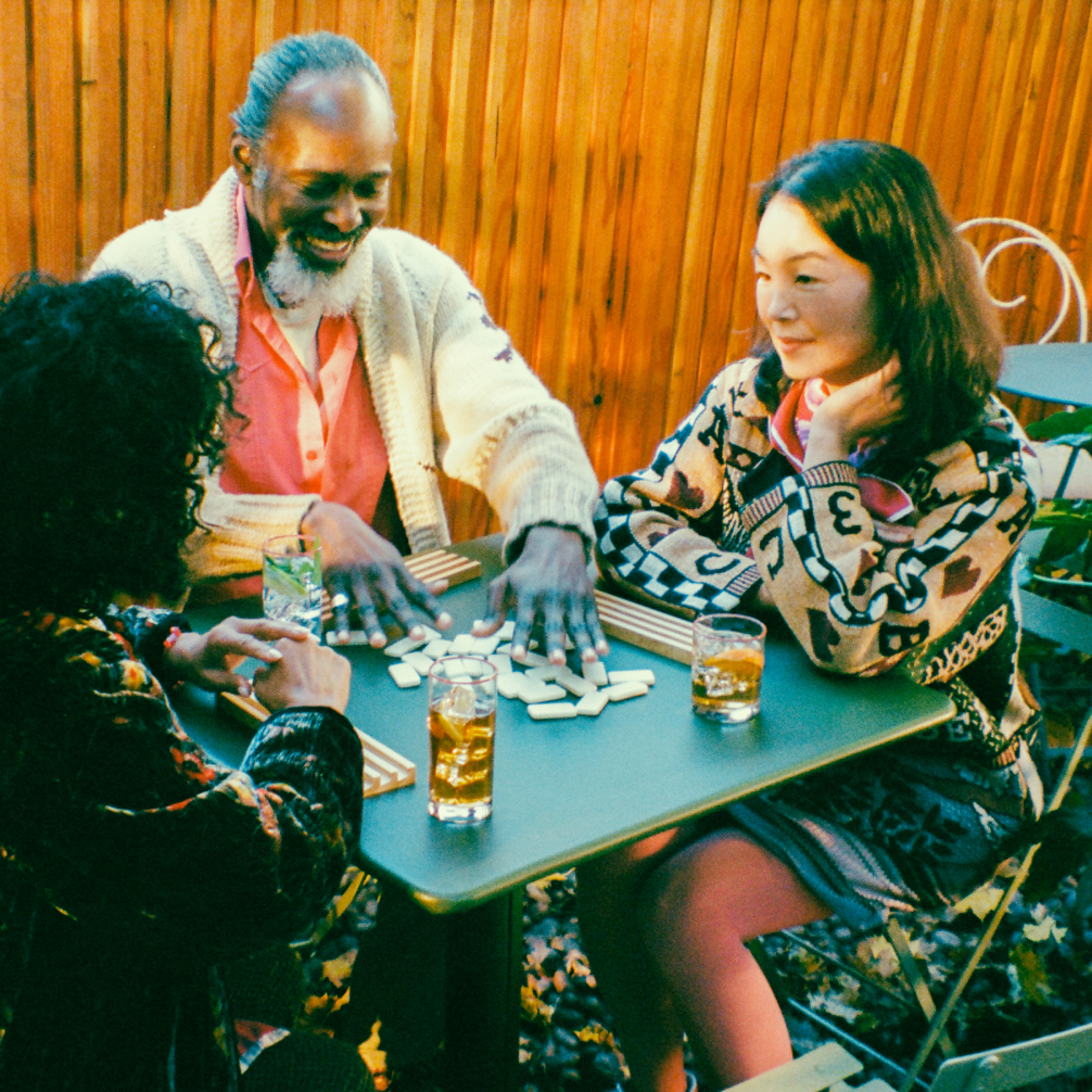 Three people sit at a table at a cafe outdoors. In front of them on the table are some dominos, which the man is shuffling, plus some drinks.