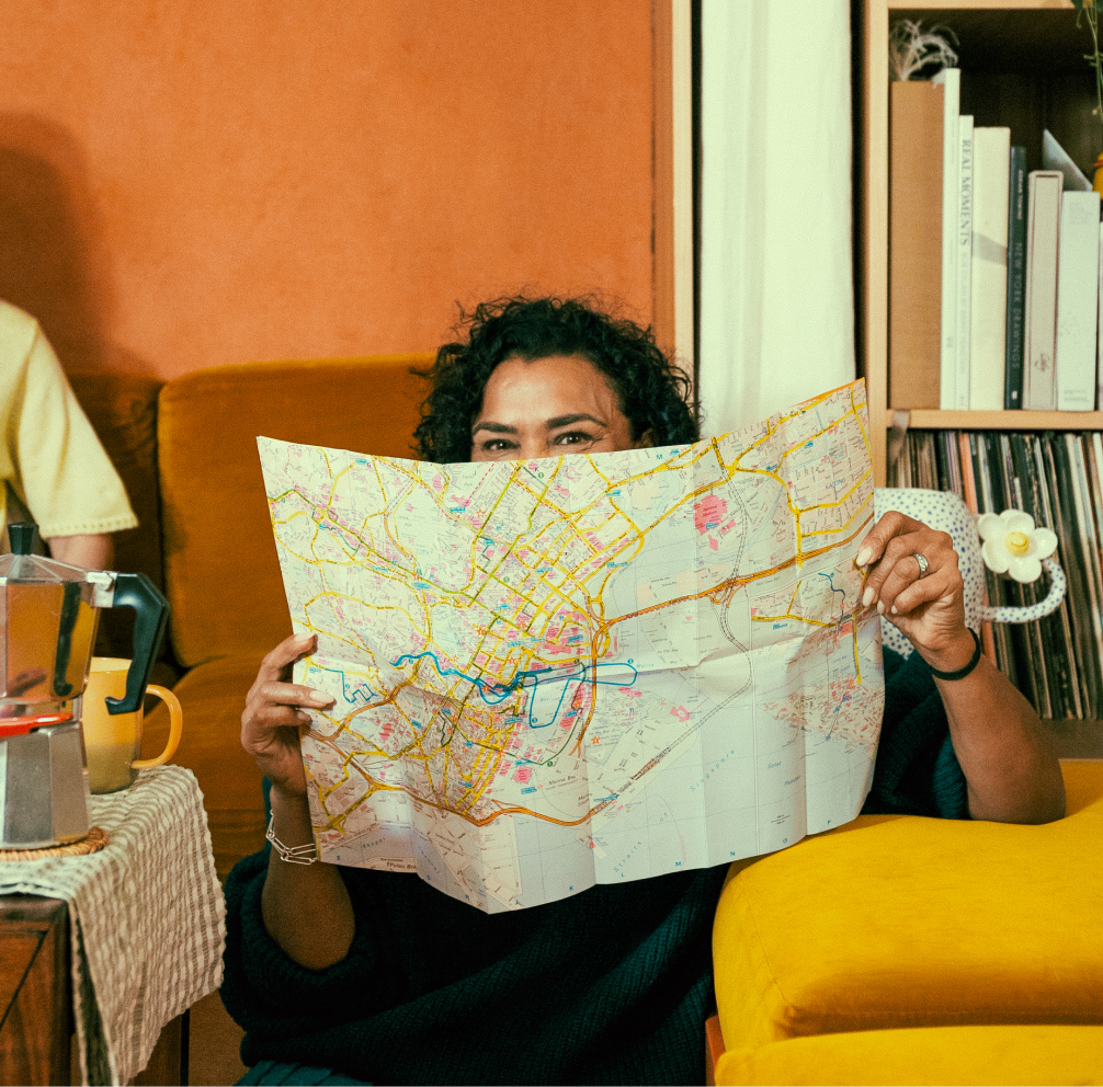 A woman sits smiling on a living room floor, facing the camera, holding up a map to obscure the lower half of her face.