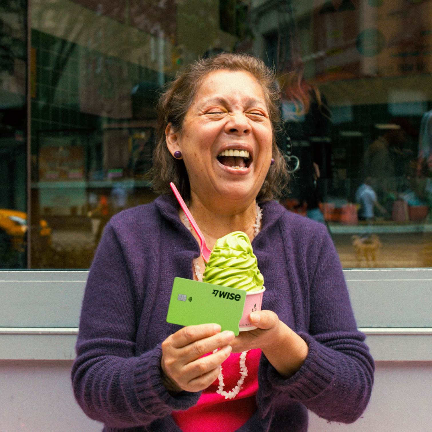 A woman stands laughing against a shop window. She holds an ice cream in one hand, and her Wise debit card in front of it in her other hand.