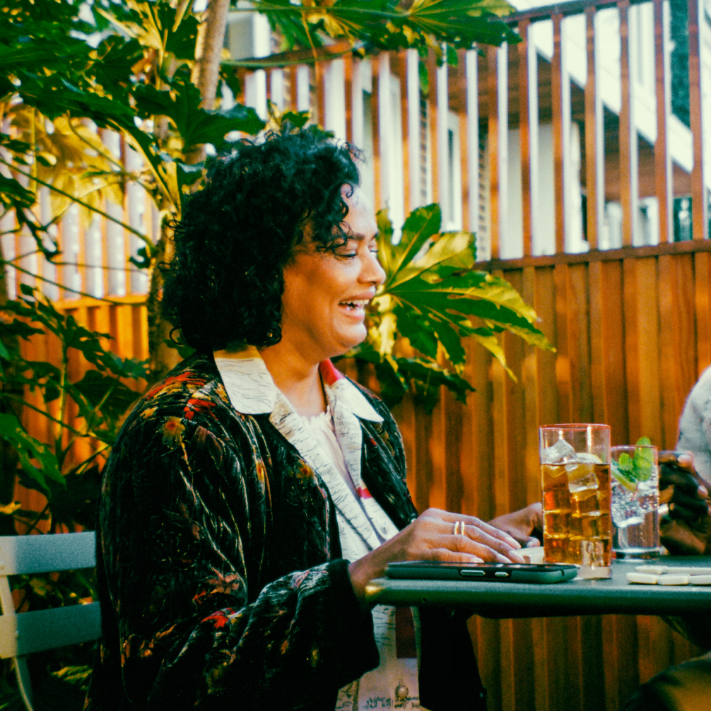 A woman sits at a table in an outdoor cafe. She laughs, facing away from the camera, with a glass of beer and some dominos on the table in front of her.