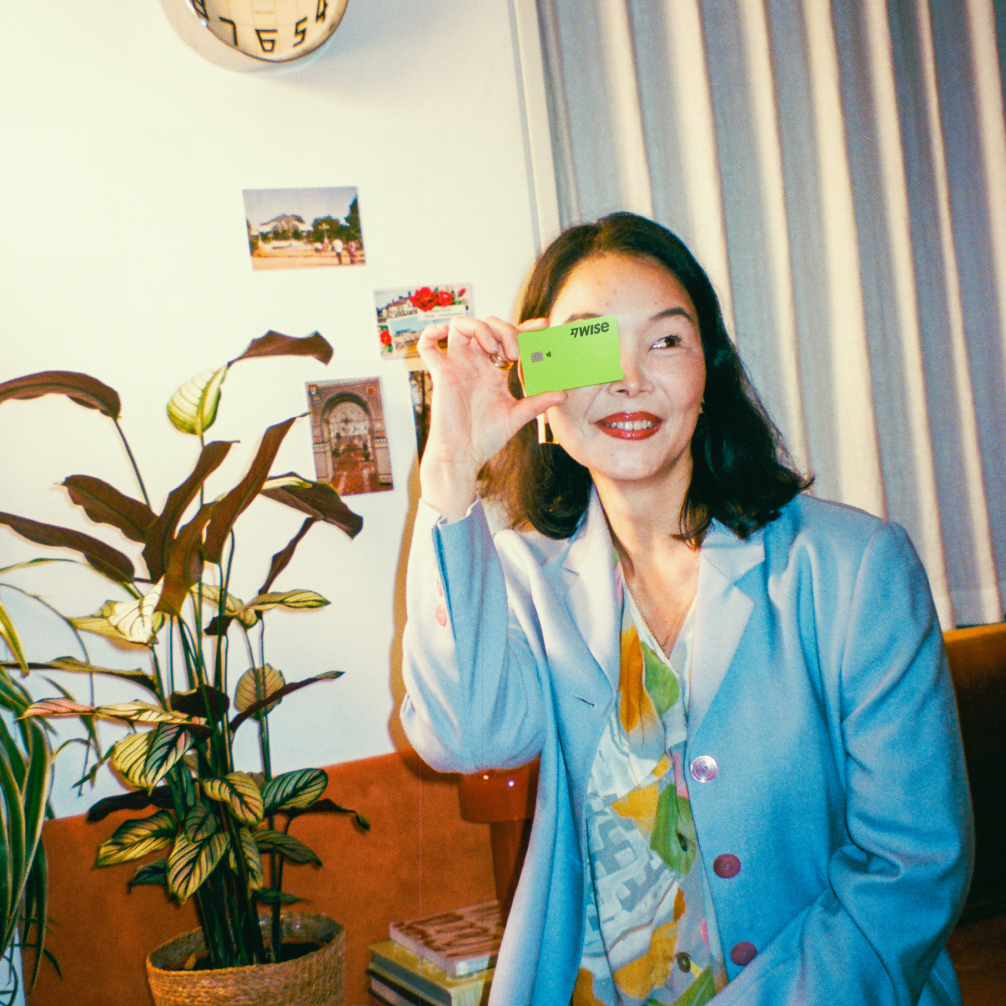 A woman sits on the corner of a chair, holding a Wise debit card over her face.