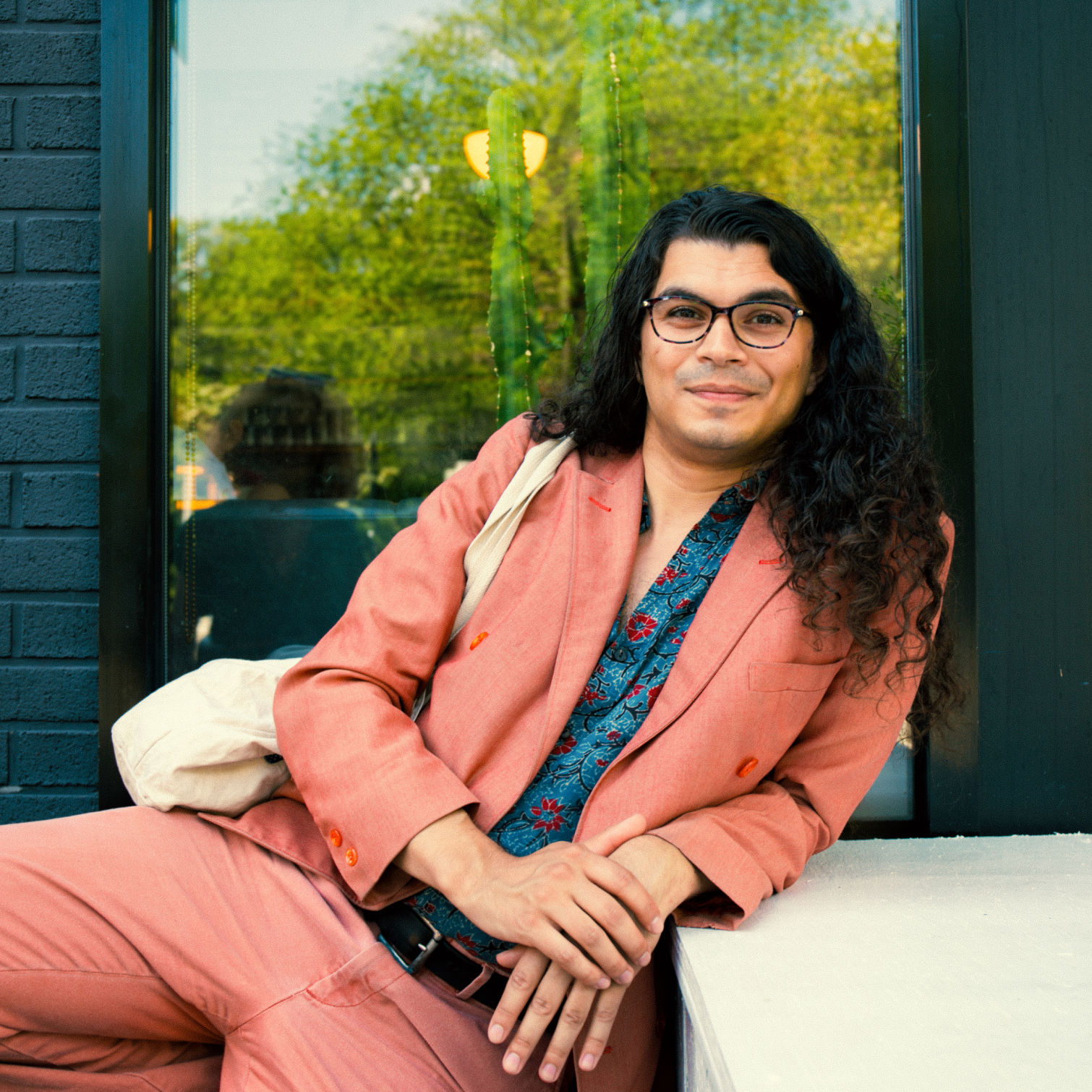 A man wearing a pink suit, sits leaning against a window and facing the camera. He rests his left arm on the table next to him, and has a tote bag slung over his right shoulder.